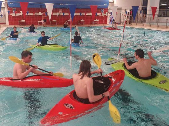 A group of three people in kayaks with others behind taking part in a kayaking session at the swimming pool